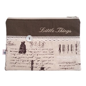 Könnte beinhalten: Eine braune Stofftasche mit Reißverschluss, weißer Spitzenborte und einem Knopf. Die Tasche ist mit beigefarbenem Stoff gefüttert und der Text "Little Things" ist auf der Vorderseite gestickt.