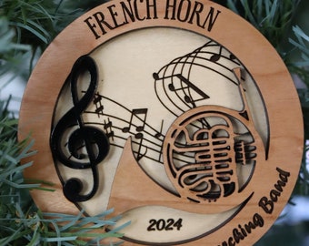 May include: A wooden Christmas ornament featuring a French horn, musical notes, and the text "French Horn Marching Band 2024".