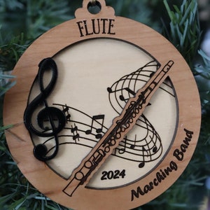 Pode incluir: Um enfeite de Natal de madeira com uma flauta, notas musicais e o texto "FLUTE Marching Band 2024".