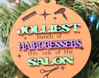 May include: Wooden Christmas ornament with the text "Jollies bunch of Hairdressers this side of the Salon." The ornament is round with a comb, hair dryer, and scissors design. The text is in green and purple.