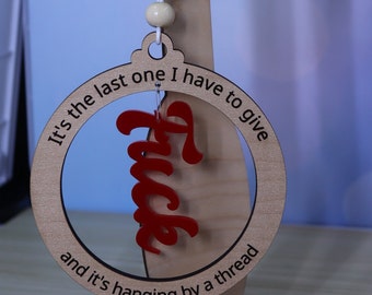 May include: A wooden Christmas ornament with the text "It's the last one I have to give and it's hanging by a thread" and the word "Fuck" written in red inside a circle.