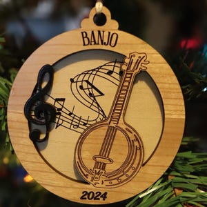 May include: Wooden banjo ornament with the word "BANJO" at the top. The ornament features a banjo illustration, musical notes, and the year "2024". The ornament is round with a light brown finish.