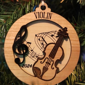 Può includere: Un ornamento natalizio in legno con un violino, note musicali e l'anno 2024 inciso su di esso. L'ornamento è etichettato "VIOLIN".