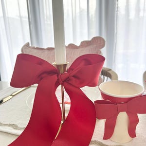 May include: A red satin ribbon tied into a large bow around a gold candle holder. The candle holder is on a white tablecloth with a white cup with a red ribbon tied around it.