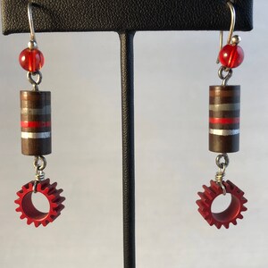 May include: A pair of earrings with red and silver accents. Each earring features a brown cylinder with silver and red stripes, a red gear, and a red bead.