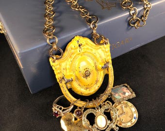 May include: A gold chain necklace with a yellow metal door handle pendant. The pendant has a heart charm, a key charm, a clock charm, and a small picture frame charm.
