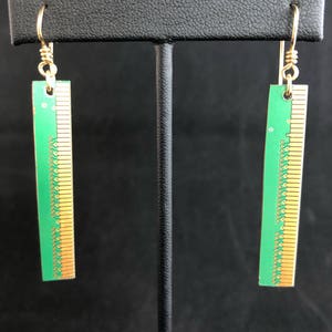 May include: A pair of unique earrings featuring rectangular green circuit board pendants with gold-colored edges and detailing. The earrings hang from gold-colored ear wires, creating a striking contrast against the dark background. These earrings are a statement piece.