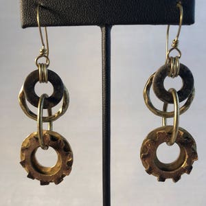 May include: A pair of gold-toned earrings with a layered design. The earrings feature two round metal pieces, one with a smooth surface and the other with a gear-like pattern.