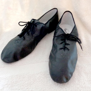 May include: A pair of black leather jazz shoes with laces. The shoes are designed for dance and have a soft, flexible sole. The shoes are shown on a white surface.