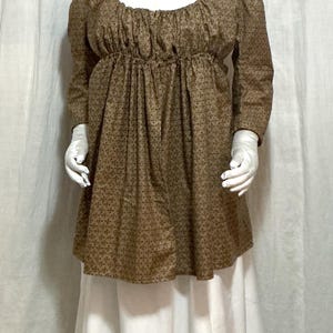 May include: A brown patterned dress with long sleeves and a square neckline. The dress has a gathered waist and is paired with a white skirt. A white floral headpiece and white gloves complete the ensemble.