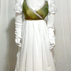 May include: A white dress with a green bodice and long sleeves is displayed on a mannequin. The bodice has a decorative trim and a wrap-style design. The dress has a high waist and a flowing skirt. The mannequin is wearing a white headpiece.