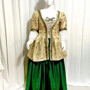 May include: A historical costume featuring a gold-colored jacket with floral embroidery and green trim, paired with a long, emerald green skirt. The jacket has short sleeves with lace cuffs. A white head covering and gloves complete the ensemble.