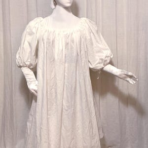 May include: A white, vintage-style dress with a round neckline and puffy sleeves. The dress is displayed on a mannequin, paired with a matching head covering and gloves. The skirt has a subtle pink trim.