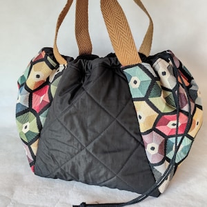 Pode incluir: Uma sacola tote preta acolchoada com fecho de cordão. As laterais apresentam um padrão geométrico colorido em vermelho, verde, amarelo e azul. A sacola tem alças tecidas marrons.