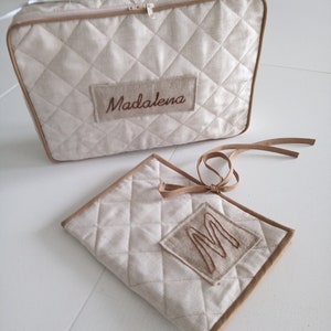 Pode incluir: Duas bolsas de tecido acolchoado bege com guarnições marrons. A bolsa maior tem um fecho de correr e uma etiqueta de nome bordada em castanho que diz "Madalena". A bolsa menor tem uma letra "M" bordada em castanho na frente.
