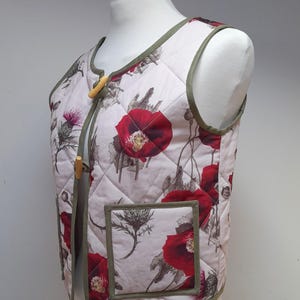 Könnte beinhalten: Gesteppte Weste mit floralem Muster aus großen roten Mohnblumen und Disteldesigns auf weißem Grund. Die Weste hat eine grüne Paspelierung und Holzschließen. Die West ist auf einer Schaufensterpuppe ausgestellt.