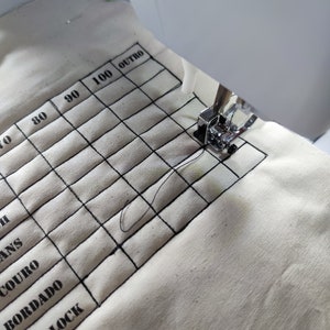 Pode incluir: Um tecido branco com um padrão de grade e os números 70, 80, 90, 100 e a palavra "OUTRO" impressos nele. O tecido está sendo costurado em uma máquina de costura.
