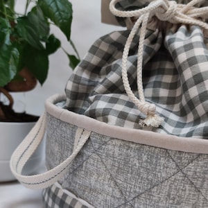 Op de afbeelding: Een grijze en witte gingham opbergmand met een witte trekkoordtas erin. De mand heeft een gewatteerde grijze buitenkant van stof en een witte rand. De tas heeft een wit trekkoord en een gingham-patroon.
