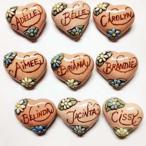 May include: Nine pink heart-shaped ceramic ornaments with floral designs and names written in brown ink. The names are: Adell, Belle, Carolyn, Aimee, Briana, Brandie, Belinda, Jacinta, and Cissy.