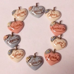May include: Ten heart-shaped ceramic pendants with different names written on them in gold lettering. Each pendant has a small flower design on the top. The pendants are in various colors, including pink, blue, and white.