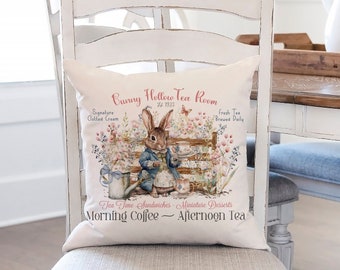 May include: A white decorative pillow with a whimsical design featuring a bunny in a blue coat, holding a teacup. The pillow has the text "Bunny Hollow Tea Room" and other text about tea and treats. The pillow is placed on a white chair.