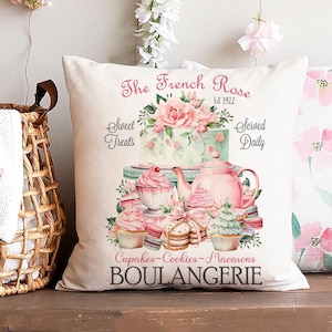 Könnte beinhalten: Ein weißes Kissen mit einer rosa und grünen Illustration einer Bäckerei namens "The French Rose Est 1922". Die Illustration zeigt eine Teekanne, Cupcakes, Kekse und Macarons. Der Text "Sweet Treats Served Daily Cupcakes-Cookies-Macarons Boulangerie" ist auf dem Kissen aufgedruckt.