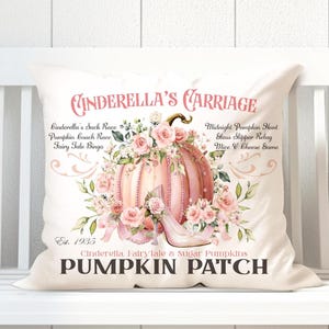 Puede incluir: Un cojín decorativo con un diseño de calabaza rosa, adornos florales y un zapato de cristal. El texto dice "Cinderella's Carriage" y "Pumpkin Patch". El cojín es de color blanco roto.