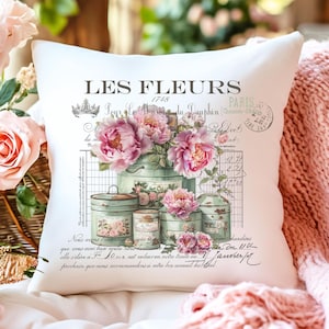 Op de afbeelding: Een wit kussen met een bloemenontwerp met roze pioenen en groene kannen. Het kussen heeft de tekst "LES FLEURS" en "1748" erop gedrukt.