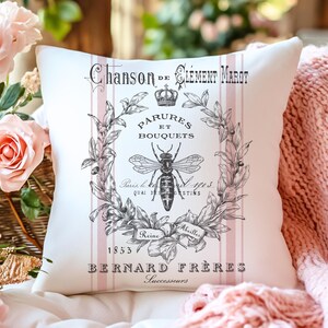 Puede incluir: Cojín cuadrado blanco con un diseño francés vintage. Presenta una ilustración en blanco y negro de una abeja, una corona y una corona floral, con texto en francés. Rayas verticales rosas acentúan el diseño. El cojín está sobre una superficie blanca.