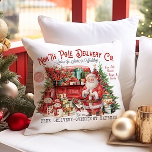 May include: A white throw pillow featuring a red truck and Santa Claus delivering presents. The truck is labelled "North Pole Delivery Co. Made in Santa's Workshop" and "Free Delivery - Overnight Express".