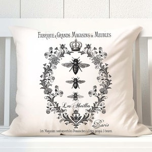 Puede incluir: Cojín cuadrado color crema con un diseño de abejas y flores negras. El cojín presenta texto en francés, incluyendo "FABRIQUE & GRANDS MAGASINS DE MEUBLES" y "Les Abeilles Paris."