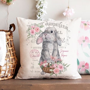 May include: A white pillow with a gray bunny holding a basket of pink flowers. The pillow has a vintage design with the text "TARDINIER" and "LES EDIT" in French.