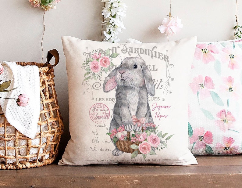 May include: A white pillow with a gray bunny holding a basket of pink flowers. The pillow has a vintage design with the text "TARDINIER" and "LES EDIT" in French.