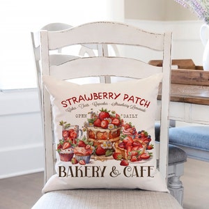 Puede incluir: Un cojín cuadrado blanco con un diseño de panadería de fresas. El cojín presenta el texto "STRAWBERRY PATCH BAKERY & CAFE" en rojo y marrón, junto con ilustraciones de pasteles, magdalenas y tartas. El cojín está sobre una silla de madera blanca.