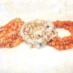 May include: Three strands of necklaces made of small, irregularly shaped orange and white beads. The necklaces are arranged in a circular pattern on a white, textured surface. The beads have a natural, organic appearance.