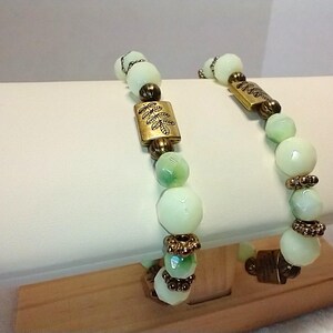 May include: Two beaded bracelets with pale green and white beads, accented with gold-colored metal details. The bracelets feature rectangular and cylindrical metal pieces with leaf designs. The bracelets are displayed on a white surface.
