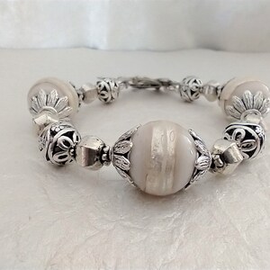May include: A beaded bracelet featuring large, light-colored, striped beads and smaller, silver-toned decorative beads. The bracelet has a silver-toned clasp and is designed with ornate metalwork. The beads are approximately 1 inch in diameter.