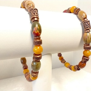 May include: Two beaded bracelets featuring a repeating pattern of smooth, multi-colored stone beads in shades of olive green, yellow, and brown, interspersed with cylindrical copper-toned metal spacers that have a textured, geometric pattern.