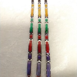 May include: Three silver bracelets with rectangular, multi-colored stone inlays. The stones are arranged in a pattern of black, yellow, green, red, and purple. The bracelets are displayed on a white surface.