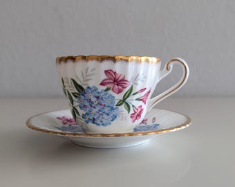 May include: White teacup and saucer set with a floral design featuring blue and pink flowers. The rim of the cup and saucer are trimmed with gold. The teacup has a curved handle.