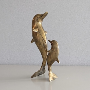 Könnte beinhalten: Zwei Delfin-Skulpturen aus Messing. Der größere Delfin steht auf seinem Schwanz, während der kleinere Delfin auf seinem Schwanz steht und hinter dem größeren Delfin positioniert ist.