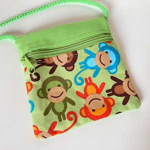 Può includere: Una piccola borsa a tracolla verde con chiusura a zip. La borsa è decorata con un motivo di scimmie di vari colori, tra cui blu, verde, arancione e marrone.