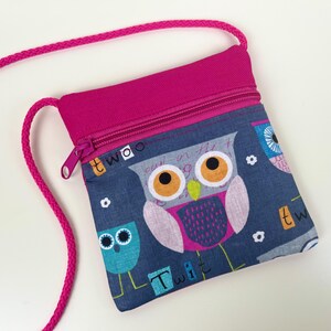Könnte beinhalten: Kleine Umhängetasche in Pink und Blau mit Reißverschluss. Die Tasche hat ein Eulenmuster in Blau, Grau, Pink und Orange. Sie hat einen pinkfarbenen Riemen und einen pinkfarbenen Reißverschluss. Die Tasche ist ca. 15 cm breit.