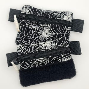 May include: Black and white zippered pouch featuring a spiderweb design. The pouch has a black zip and a black strap. The bottom of the pouch has a black, fluffy material. Designed for carrying small items.