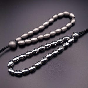 Op de afbeelding: Twee kralenaccessoires. Eén is matgrijs van kleur en de andere is glanzend zilver van kleur. Beide hebben een ovaal kralenontwerp en zijn geregen op een zwart koord. De accessoires worden op een zwart oppervlak weergegeven.