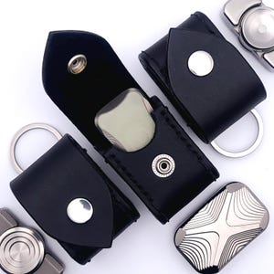 May include: Black leather cases with silver snap closures and key rings. One case is open, revealing a light-colored, rounded object. Several silver fidget spinners are also visible, showcasing a modern design.