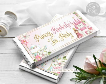 Könnte beinhalten: Zwei rechteckige Schokoriegelverpackungen mit einem rosa-weißen Blumenmuster und goldenem Schriftzug, der Princess Kimberly's Tea Party lautet. Die Grafiken zeigen ein kleines Schloss, eine Teekanne und eine Prinzessin.