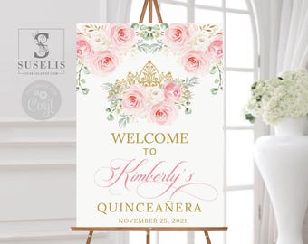 May include: A rectangular welcome sign featuring a gold crown graphic centered above pink and white watercolor roses and green leaves. The text reads: WELCOME TO Kimberly's QUINCEAÑERA NOVEMBER 25, 2021 in gold and pink script and block lettering.