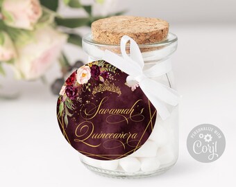 May include: A clear glass jar filled with white candies, sealed with a cork stopper. A round tag is tied to the neck with a white ribbon, featuring a dark burgundy watercolor background, gold floral accents, a gold crown, and the text Savannah's Quinceañera.