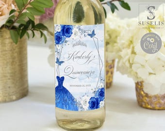 May include: A clear glass wine bottle with a decorative label featuring blue roses, a blue ballgown, a silver tiara, and butterflies. The text reads Kimberly's Quinceañera and NOVEMBER 25, 2023. The design includes a geometric frame on a light blue watercolor background.